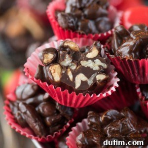 Close-up of glossy nut clusters of dark chocolate candy with nuts, placed in red and pink paper cups, stacked on a plate. Various nut pieces are visible inside the rich chocolate.