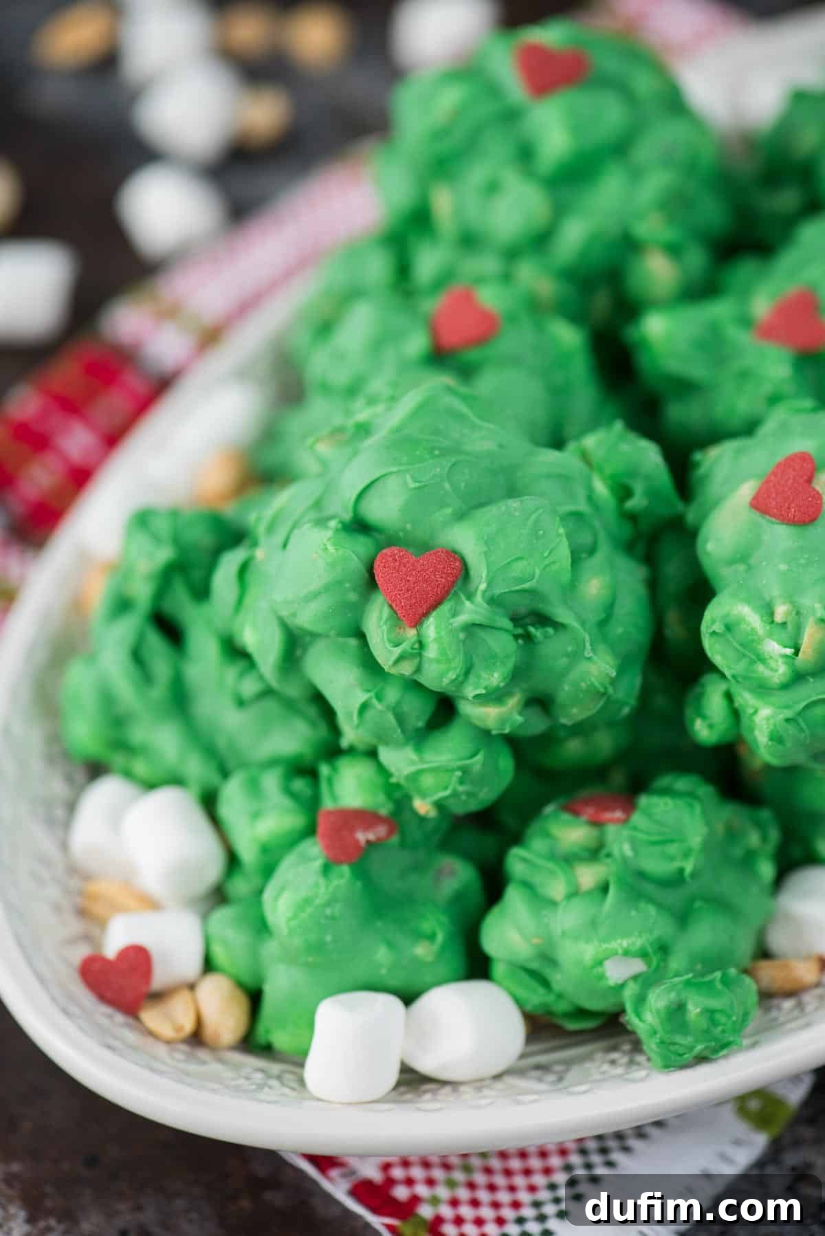A hand placing a red heart sprinkle on a freshly scooped green Grinch marshmallow cluster, surrounded by more finished clusters.