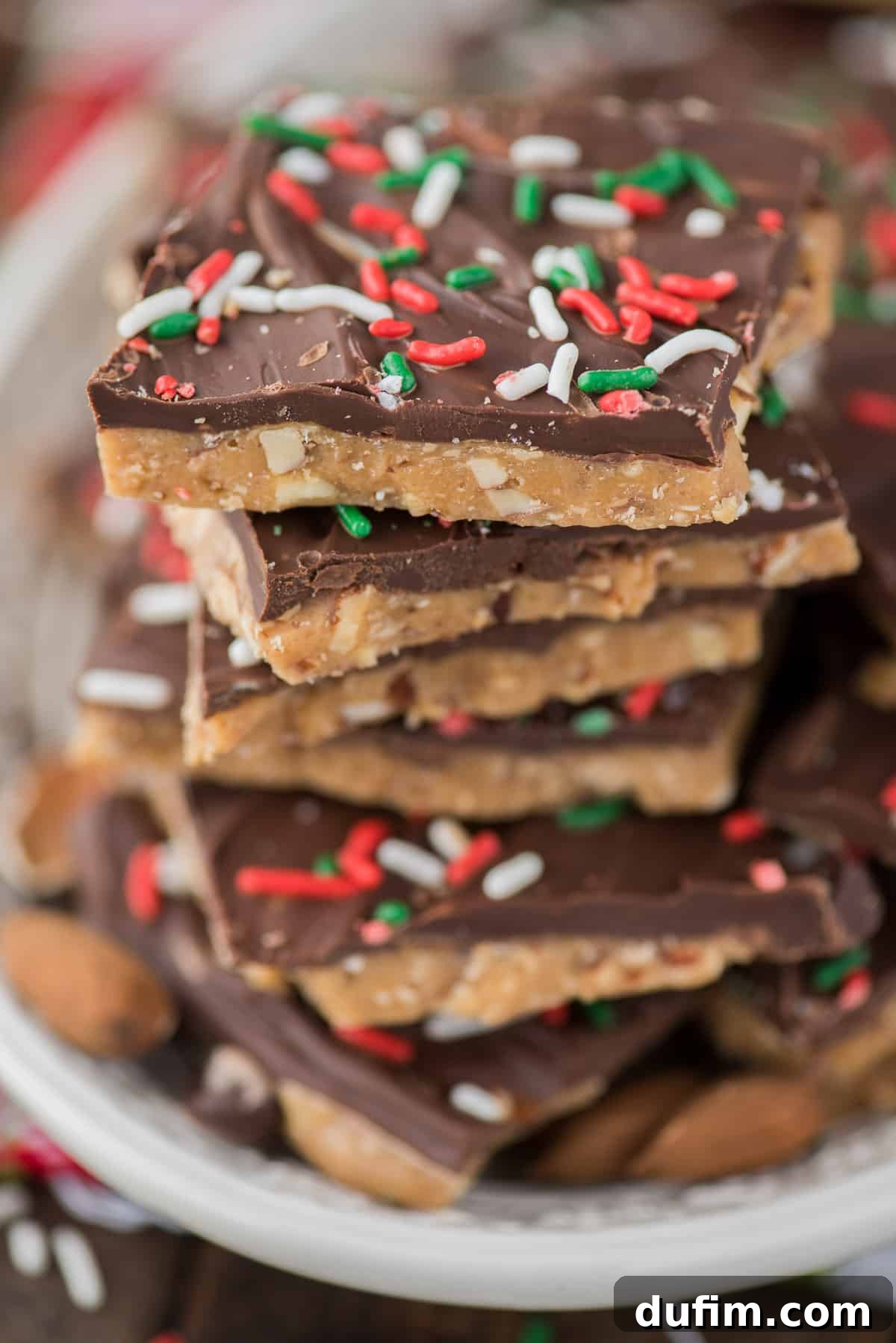 Stacked pieces of golden English toffee adorned with melted chocolate and festive Christmas sprinkles, resting on a white plate.