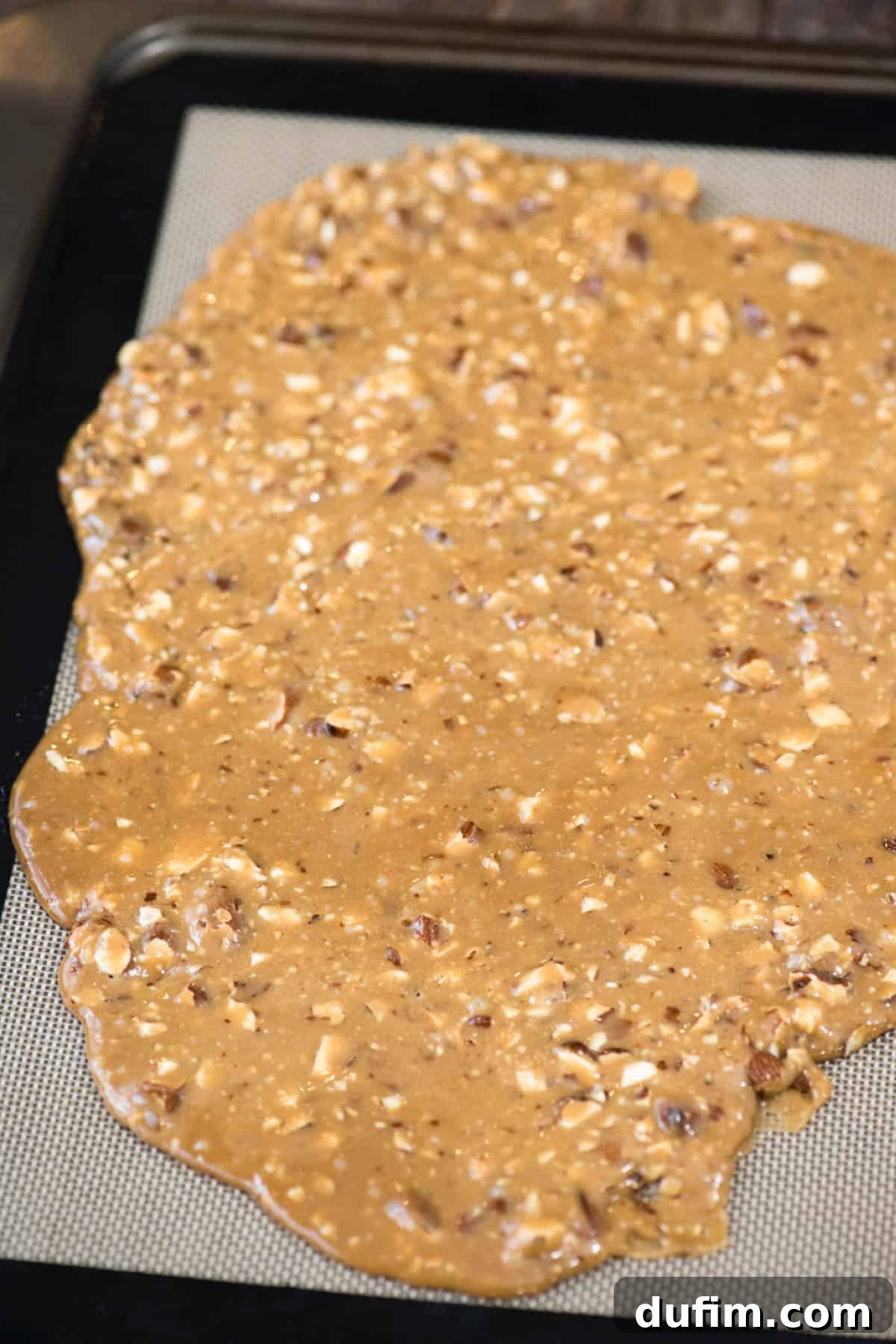 Golden, freshly poured toffee spread evenly across a white silicone baking mat, showing its smooth texture before chocolate is added.