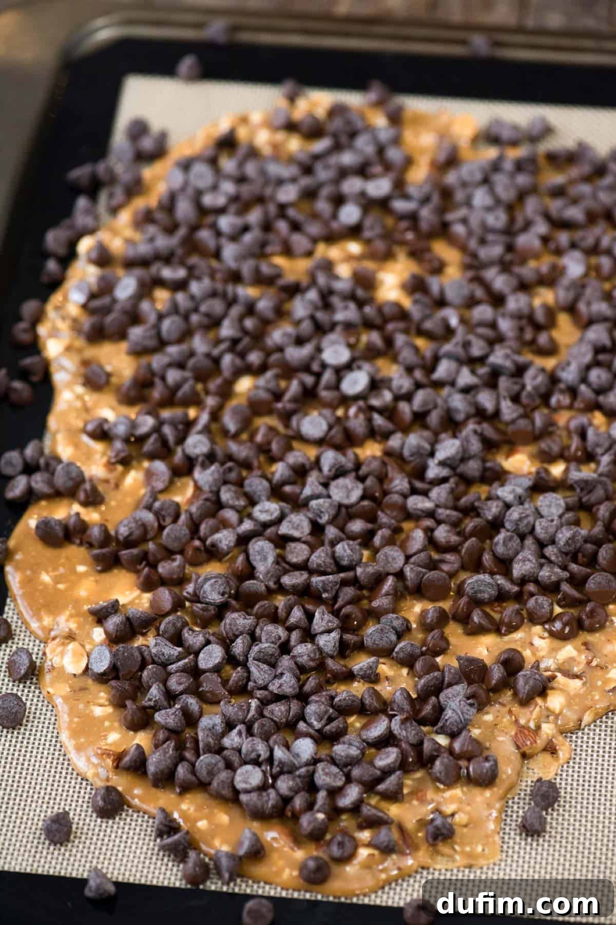 Toffee spread across a silicone baking mat, topped with a generous layer of milk chocolate chips beginning to melt from the residual heat.