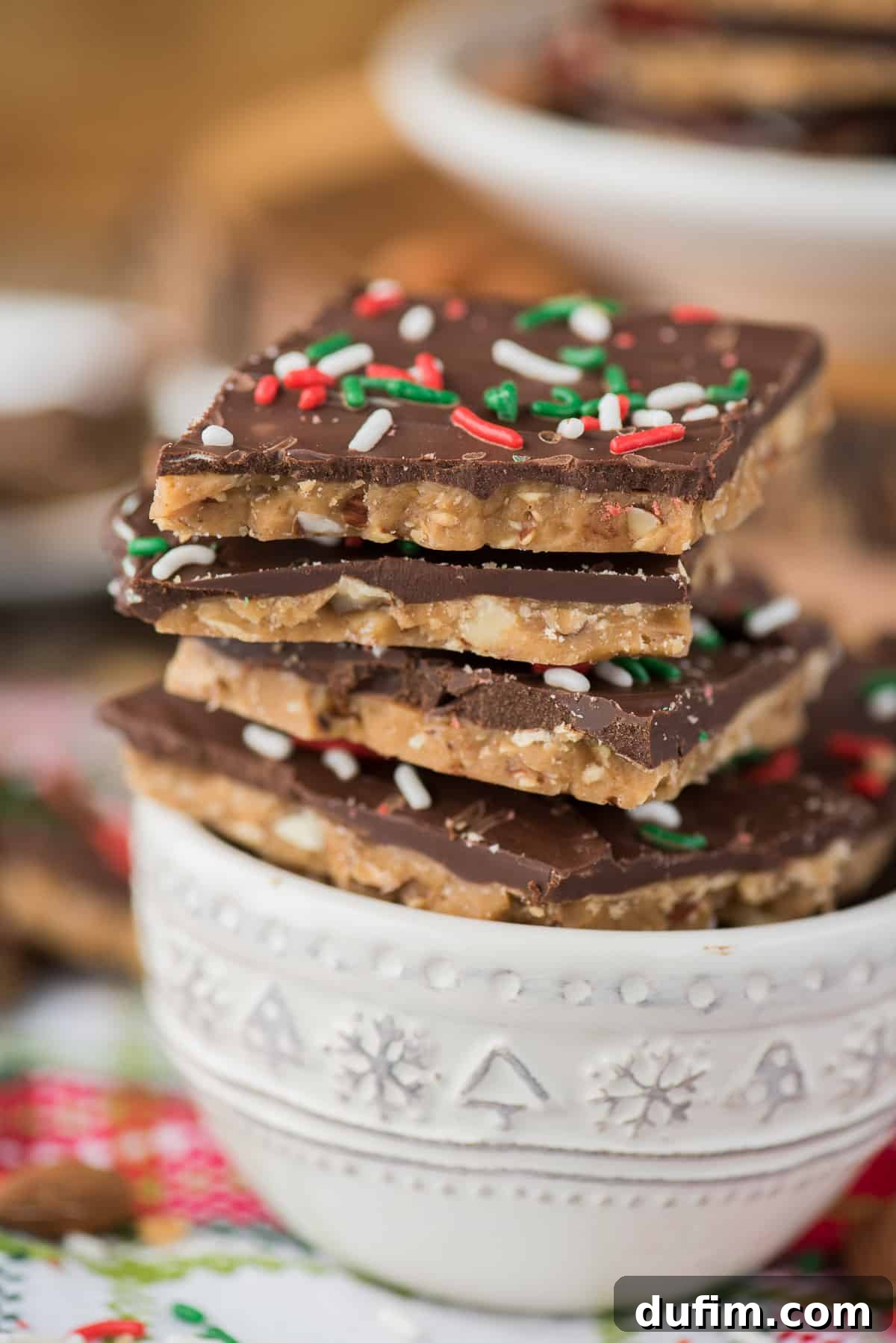 Several pieces of finished English toffee, topped with chocolate and colorful Christmas sprinkles, neatly arranged in a white bowl.