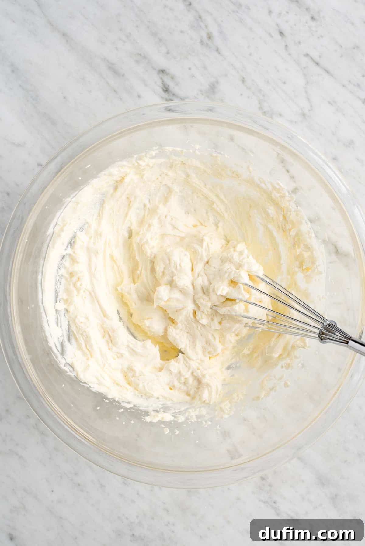 Silky smooth cheesecake flavored whipped cream in a clear glass bowl with a whisk resting inside, showcasing its creamy texture.