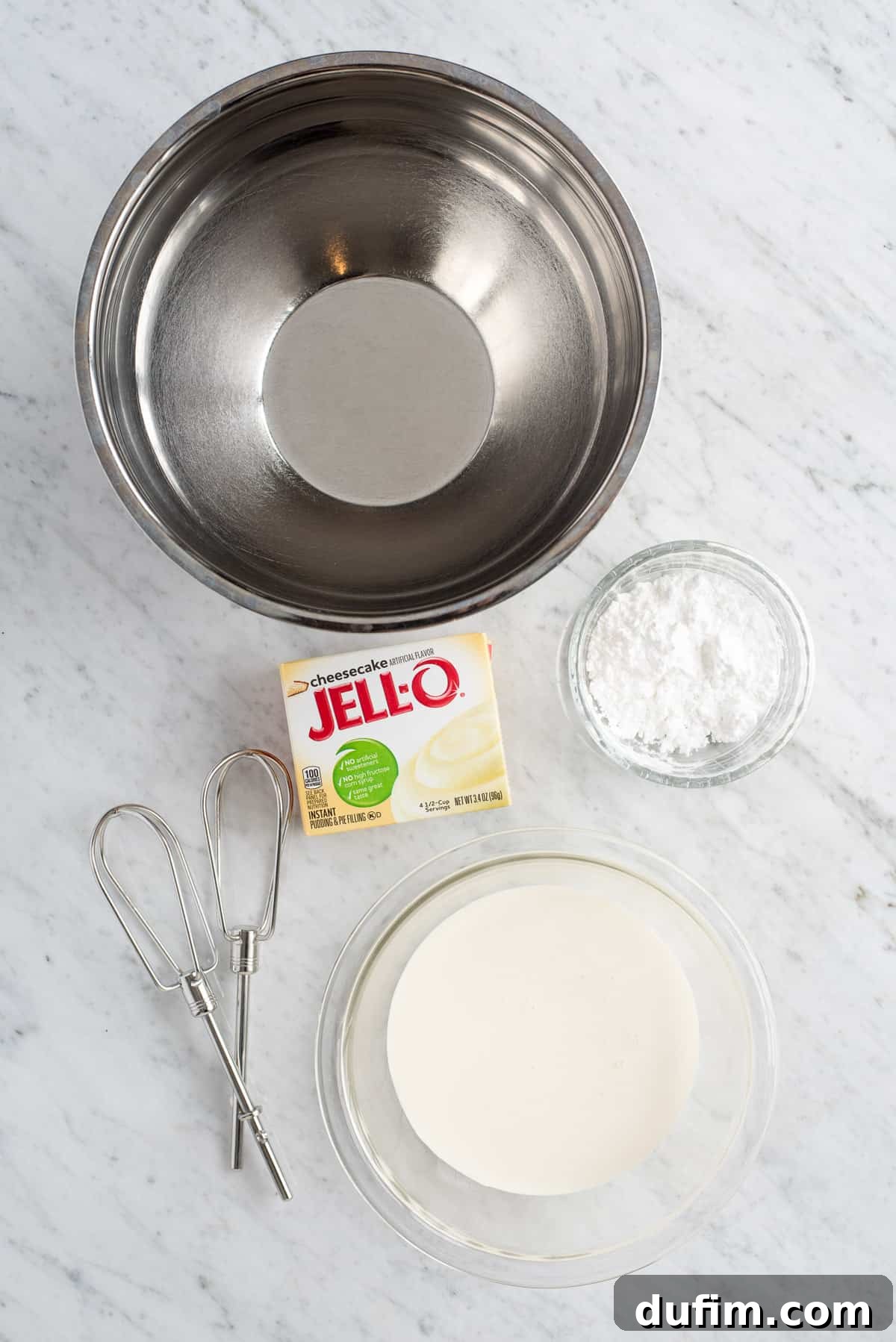 Ingredients for making cheesecake whipped cream displayed: a metal mixing bowl, powdered sugar, heavy whipping cream, and a box of cheesecake flavored instant pudding mix, alongside electric mixer beaters.