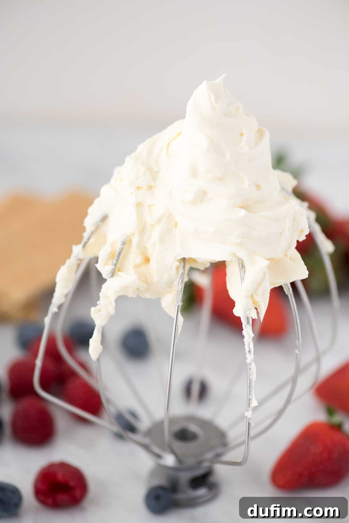 Close-up of a KitchenAid whisk attachment coated with perfectly whipped cheesecake flavored cream, with fresh berries blurred in the background.