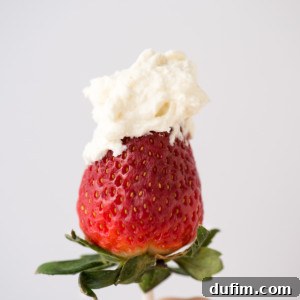 A hand holding a fresh strawberry on a stick, topped with a generous dollop of cheesecake whipped cream against a plain light background.
