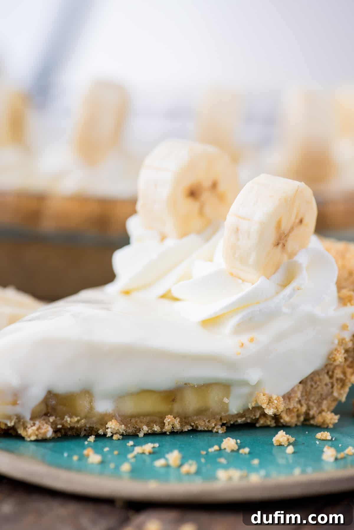 A perfect slice of creamy banana breeze pie topped with elegant whipped cream swirls and a fresh banana slice, presented on a vibrant teal plate.