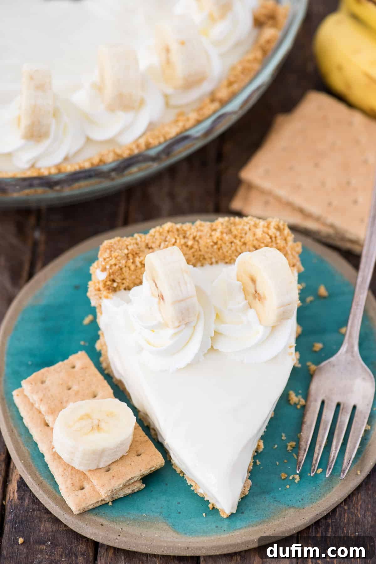 A pristine slice of Banana Breeze Pie, featuring layers of graham cracker crust, banana, creamy filling, and whipped cream, elegantly plated on a teal dish.