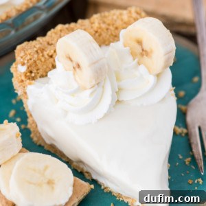 A slice of creamy banana pie on a blue plate, topped with whipped cream and banana slices, with extra banana slices and a fork beside it.