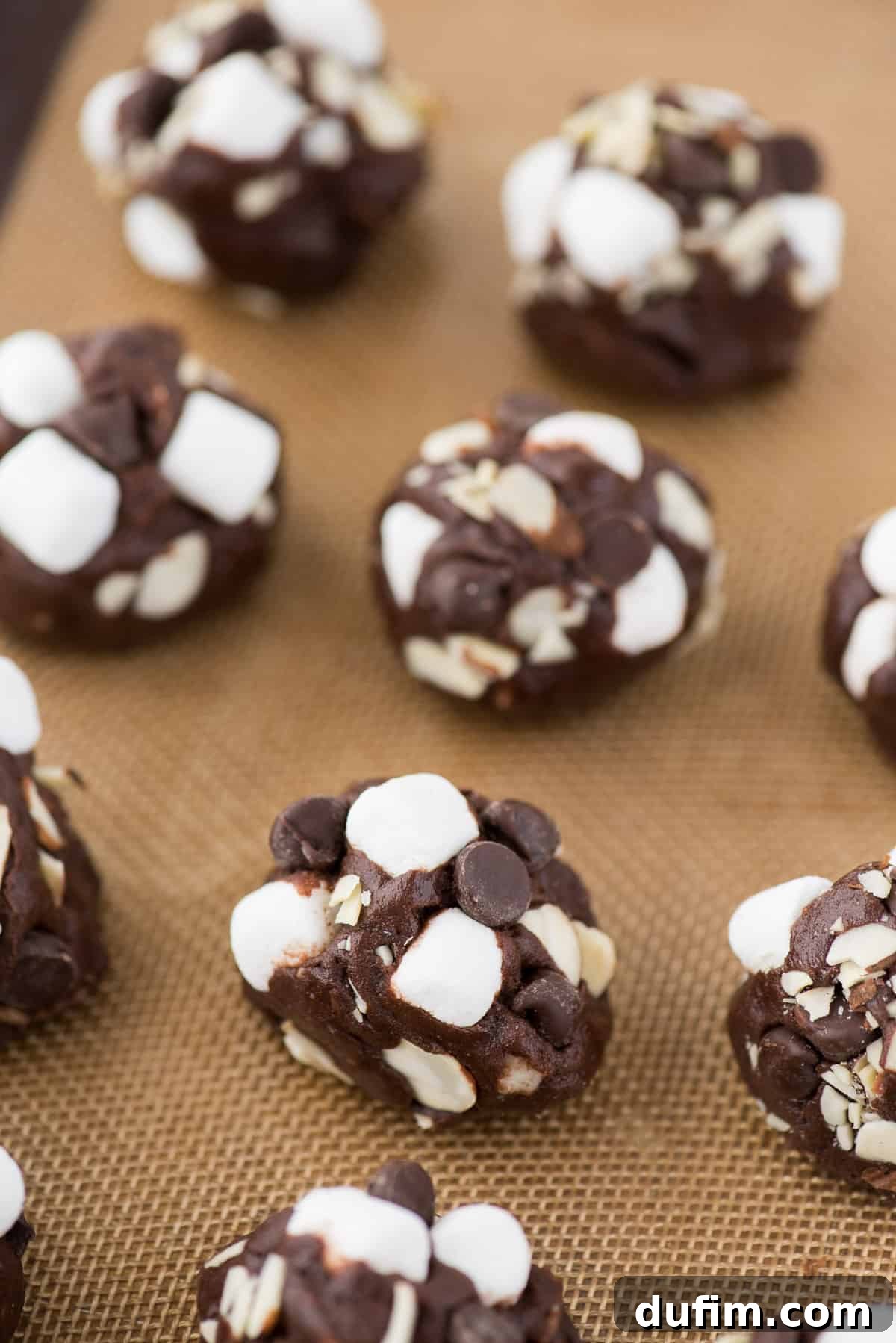 Soft and chewy rocky road cookies will hit the spot and remind you of your favorite rocky road ice cream. These chocolate cookies are packed with almonds, chocolate chips and marshmallows! You don’t have to chill the dough, this rocky road cookie recipe is ready in 30 minutes! Scooped rocky road cookie dough balls, generously topped with almonds, chocolate chips, and mini marshmallows, ready for baking on a silicone mat.