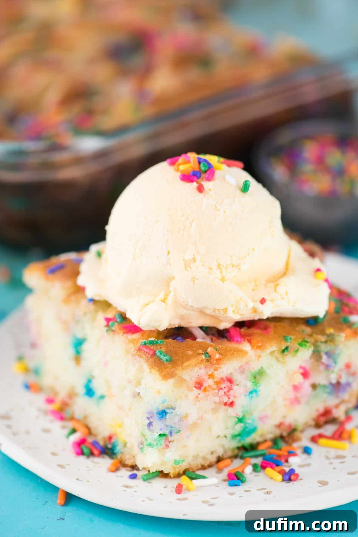 A close-up of a Funfetti Earthquake Cake slice, adorned with a melting scoop of vanilla ice cream and scattered colorful sprinkles, presented on a white plate on a blue surface.