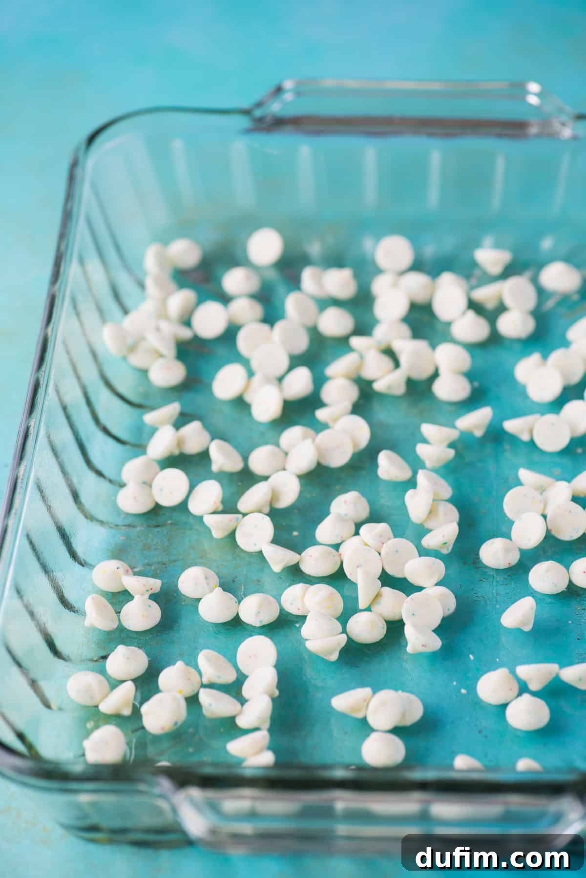 A glass 9x13 inch baking pan with a generous layer of white confetti chocolate chips evenly spread across the bottom.