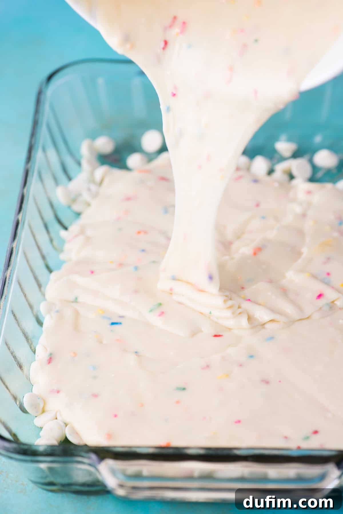 Light pink funfetti cake batter, studded with colorful sprinkles, being poured gently into a glass 9x13 inch baking pan already containing a layer of confetti chocolate chips.