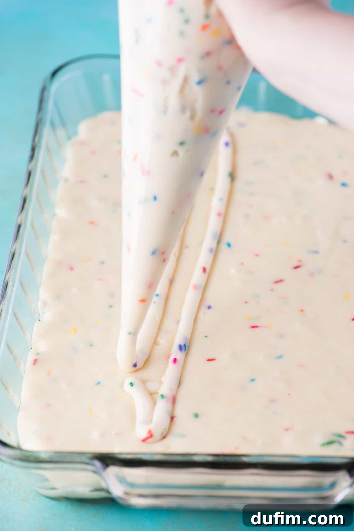 Cream cheese frosting being piped in a grid pattern over the funfetti cake batter in a glass 9x13 inch baking pan.