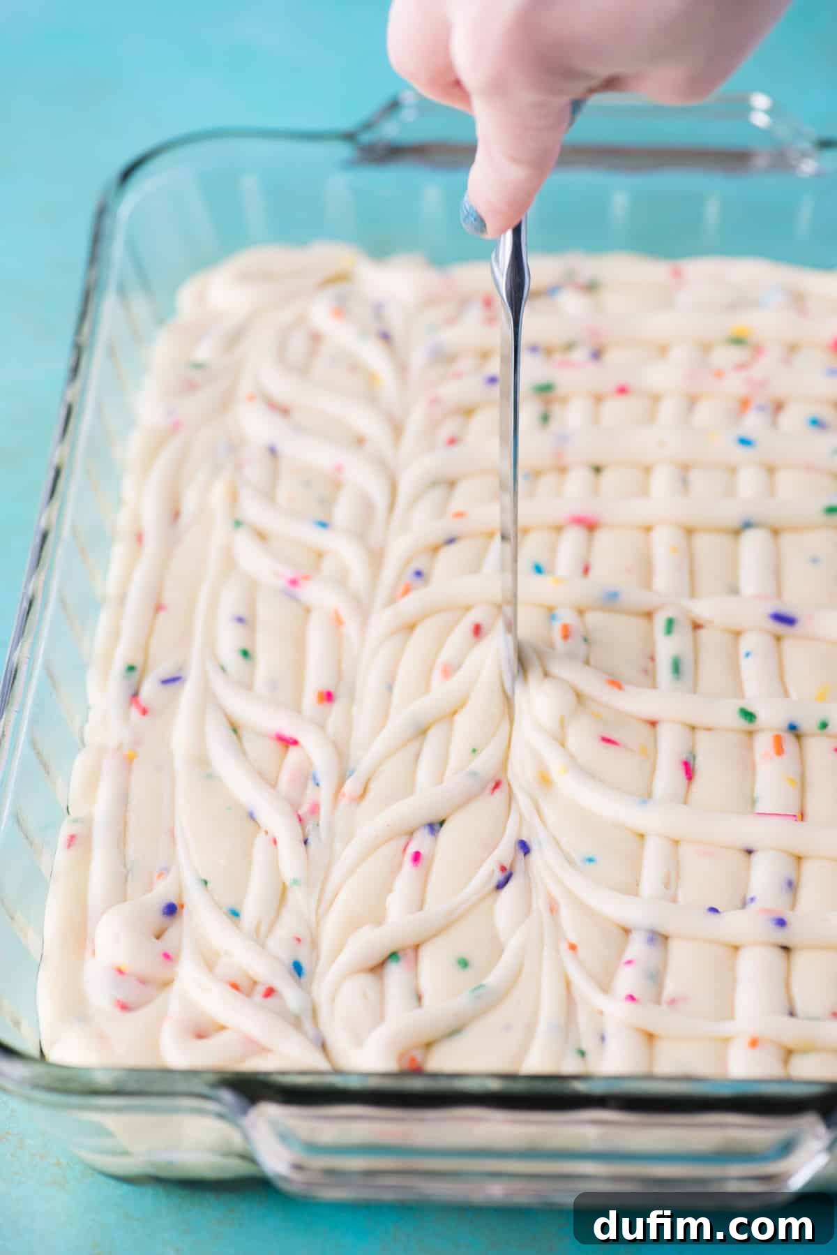 A butter knife is used to gently swirl the piped cream cheese frosting and funfetti cake batter together in a glass 9x13 inch pan, creating marbling patterns.