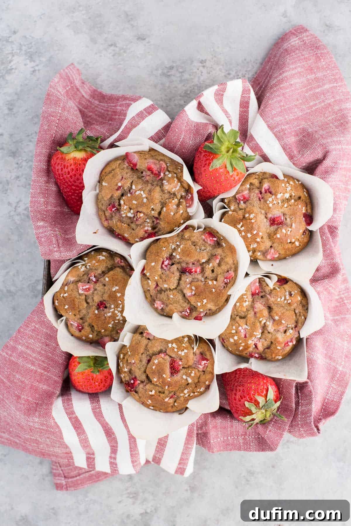 A charming stack of golden-brown strawberry banana muffins, nestled in white parchment paper tulip liners, resting gracefully on a festive red and white striped towel.