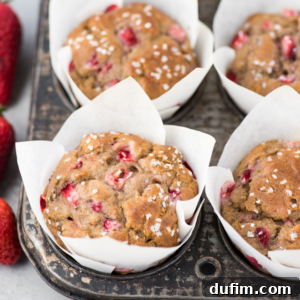 Four freshly baked strawberry banana muffins in a metal tin, each beautifully wrapped in white parchment paper tulip liners. The muffins are delicately topped with coarse sugar and visible chunks of fresh strawberries. A scattering of fresh whole strawberries completes the scene.