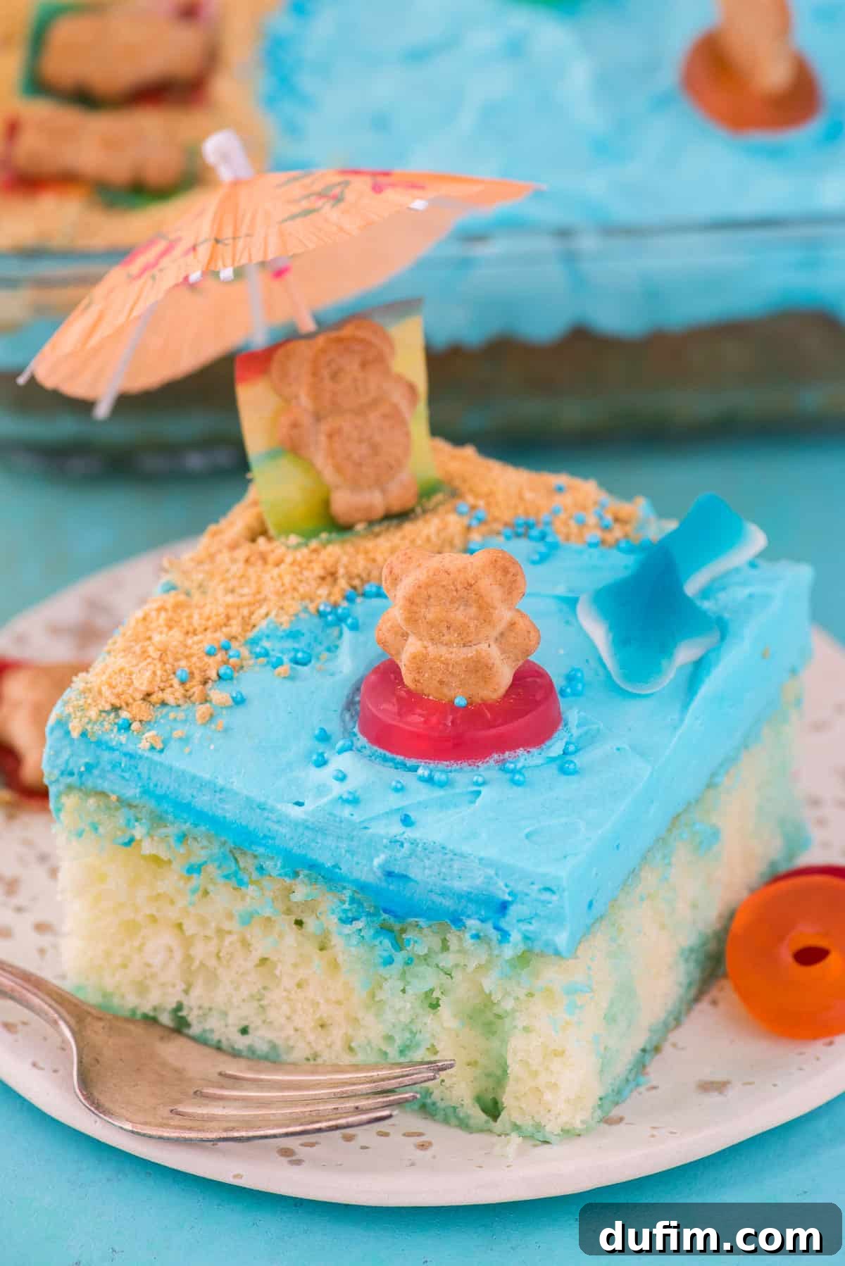Delicious Beach Poke Cake Slice for a Beach Party A beautifully decorated slice of beach theme poke cake on a white plate, showing layers of moist cake, blue jello, and creamy frosting topped with graham cracker sand, gummy shark, and a teddy graham on a fruit roll-up towel.