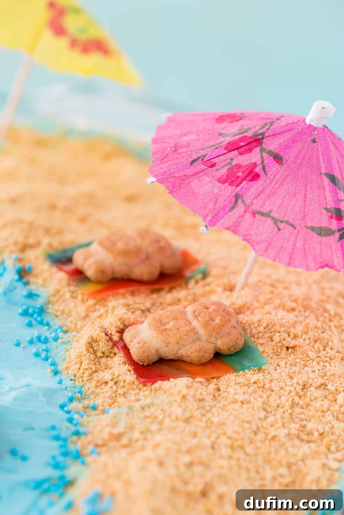 Creative Beach Cake Decorations with Teddy Grahams Overhead view of a beautifully decorated beach theme cake, featuring teddy grahams on fruit roll-up towels, graham cracker sand, and a mini toothpick umbrella for a realistic beach scene.