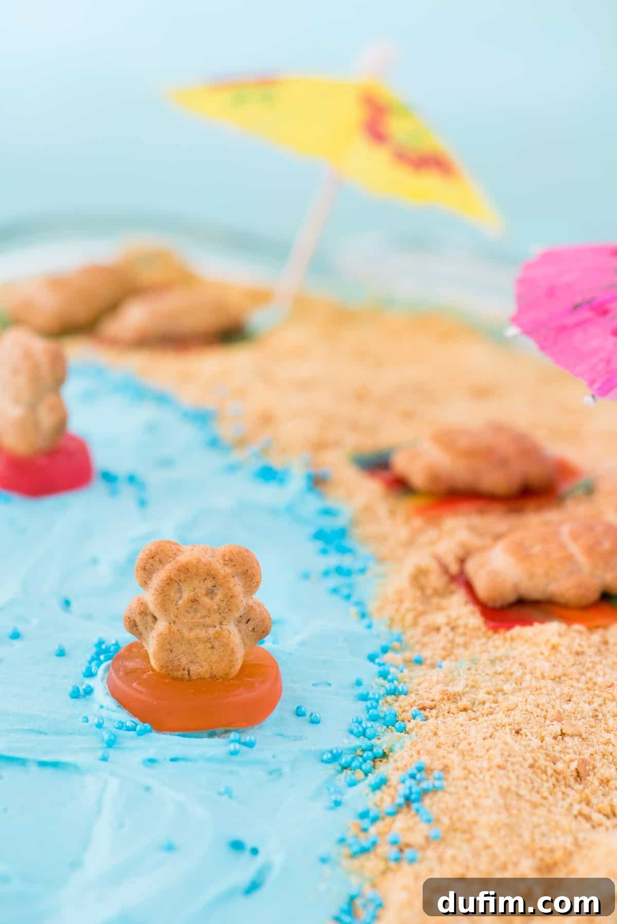 Adorable Teddy Graham Swimming Decoration on Beach Cake Close-up of a beach theme cake decoration showing a teddy graham 'swimming' in a gummy life saver in the blue jello part of the cake, with graham cracker sand nearby.