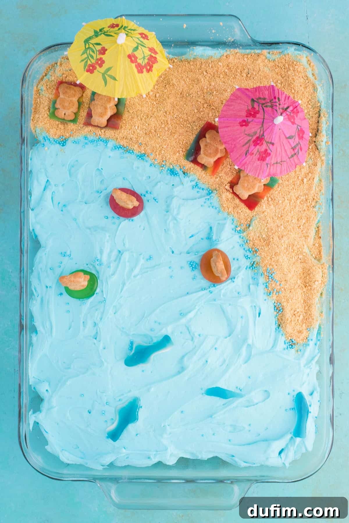 Aerial View: Beach Poke Cake for Summer Celebrations Stunning aerial photograph of a beautifully decorated beach theme poke cake, showcasing the blue jello ocean, graham cracker sand, and intricate candy decorations against a serene blue background.