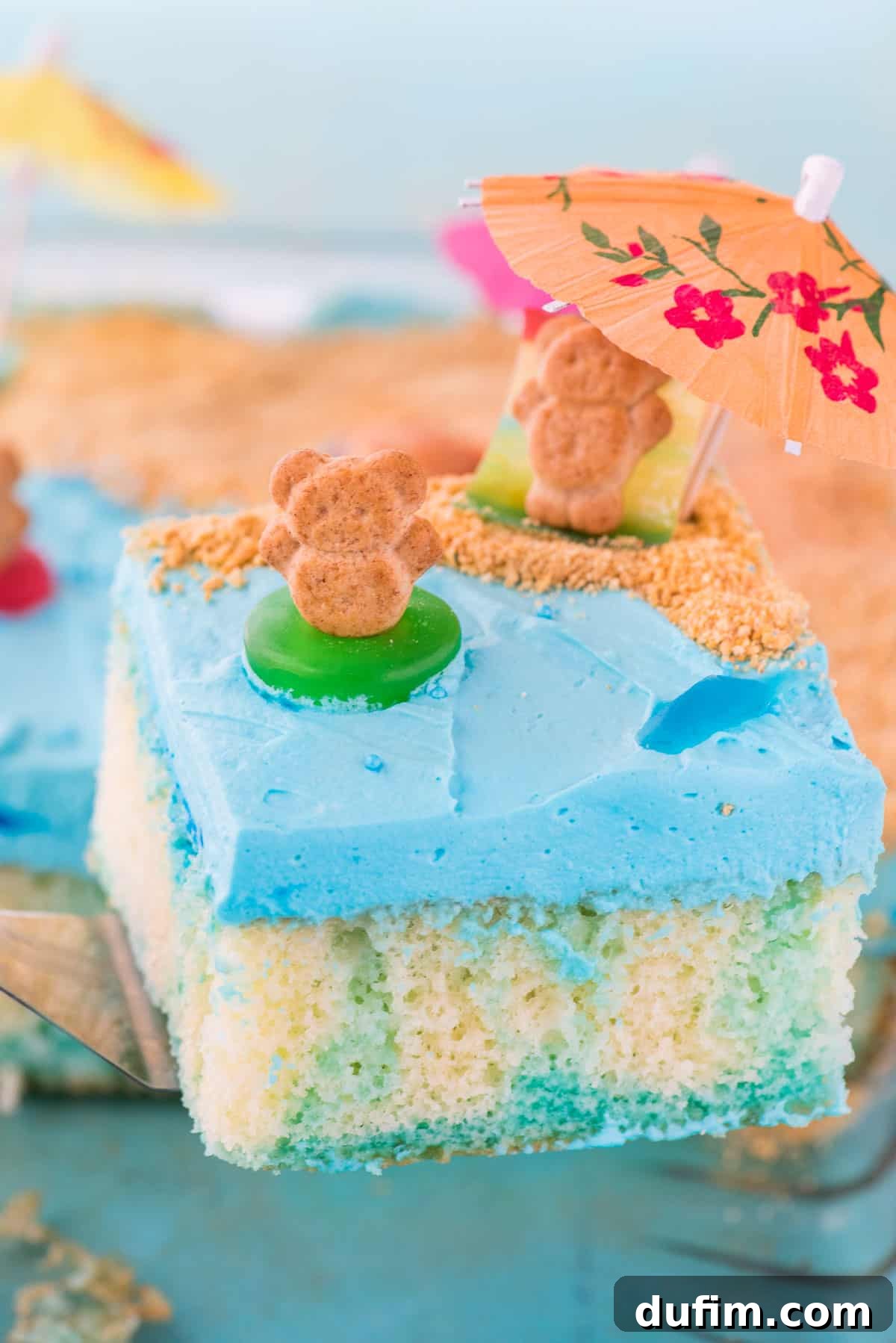 A Delicious Slice of Beach Poke Cake A vibrant slice of beach theme poke cake, beautifully layered with moist cake, blue jello, creamy frosting, graham cracker sand, and charming gummy sea creatures.