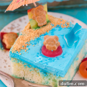 A beach poke cake slice with blue frosting, cookie crumbs for sand, teddy bear-shaped cookies, a gummy shark, and a mini paper umbrella—decorated to resemble a sweet beach scene.