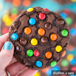 A hand with sparkly blue nail polish holds a large chocolate M&M cookie topped with colorful candies and chocolate chips. In the blurred background, more delicious chocolate M&M cookies can be seen.