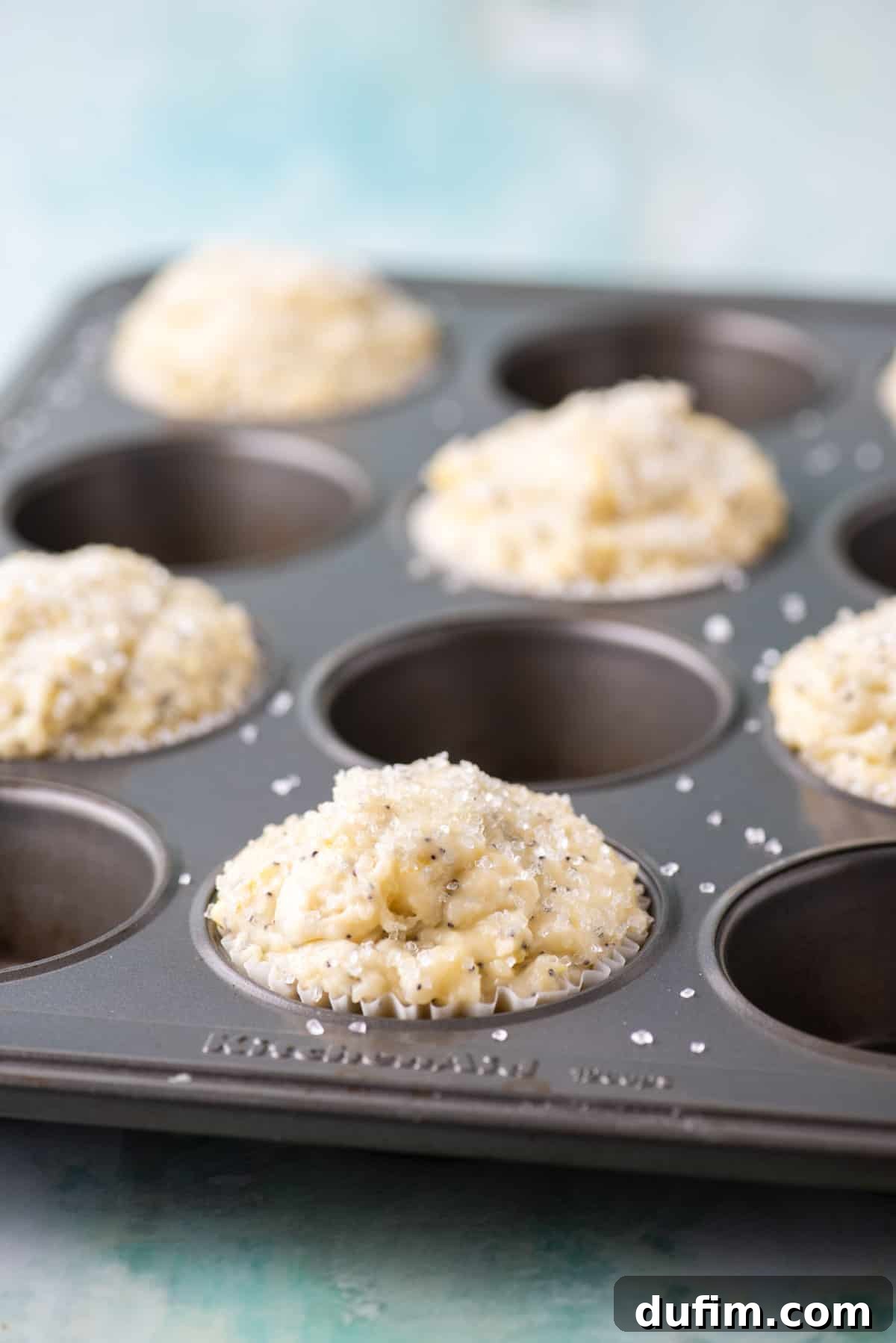 Zesty Lemon Poppy Seed Delights 6 Close-up of thick lemon poppy seed muffin batter generously scooped into a muffin pan, with every other cup filled high to allow for maximum dome growth during baking. Sanding sugar glistens on the tops.