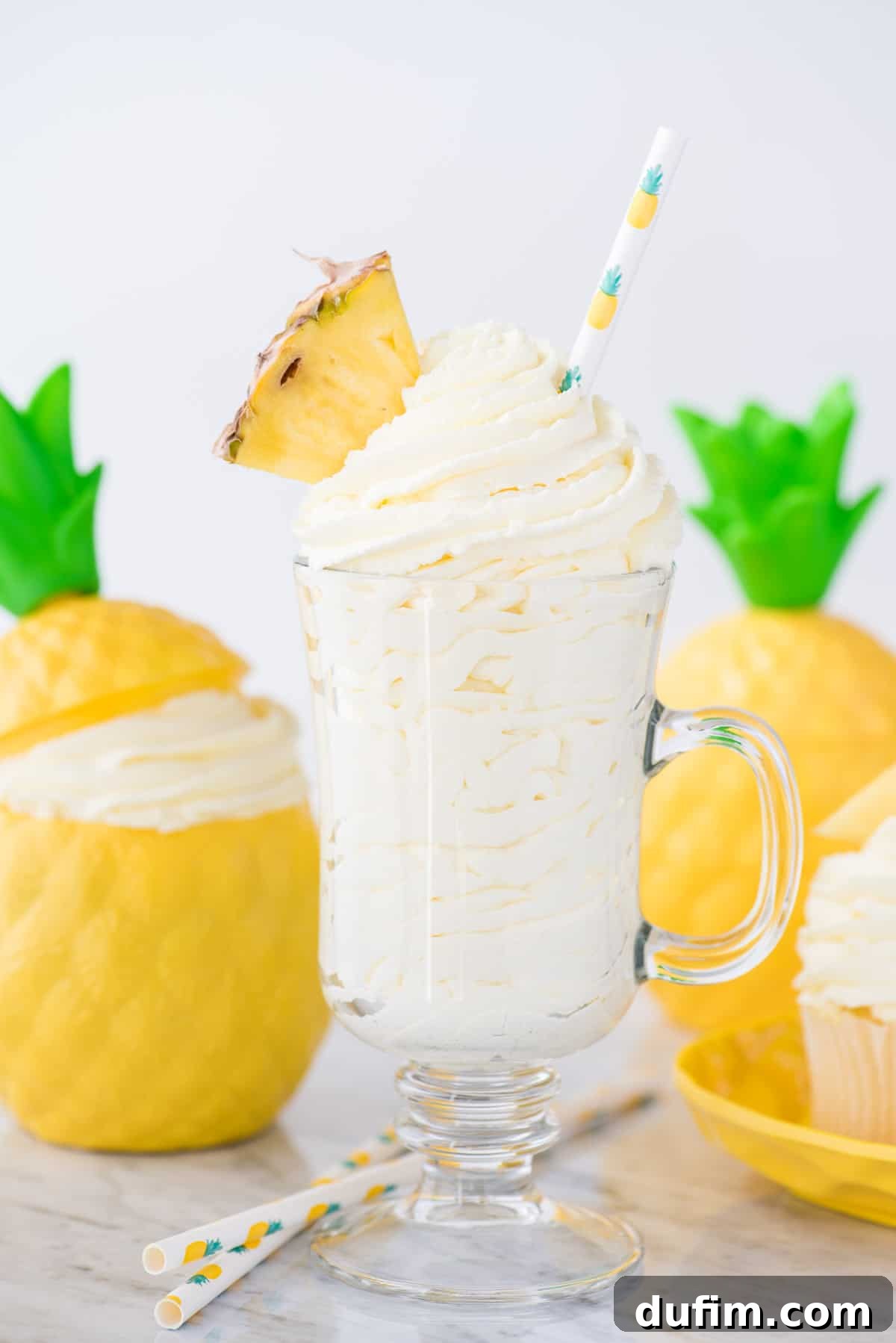 A beautiful glass cup filled with creamy pineapple whipped cream, garnished with a fresh pineapple wedge and a charming pineapple-printed straw, set against a vibrant tropical background. This image captures the essence of a refreshing, easy-to-make dessert, ready to be enjoyed.