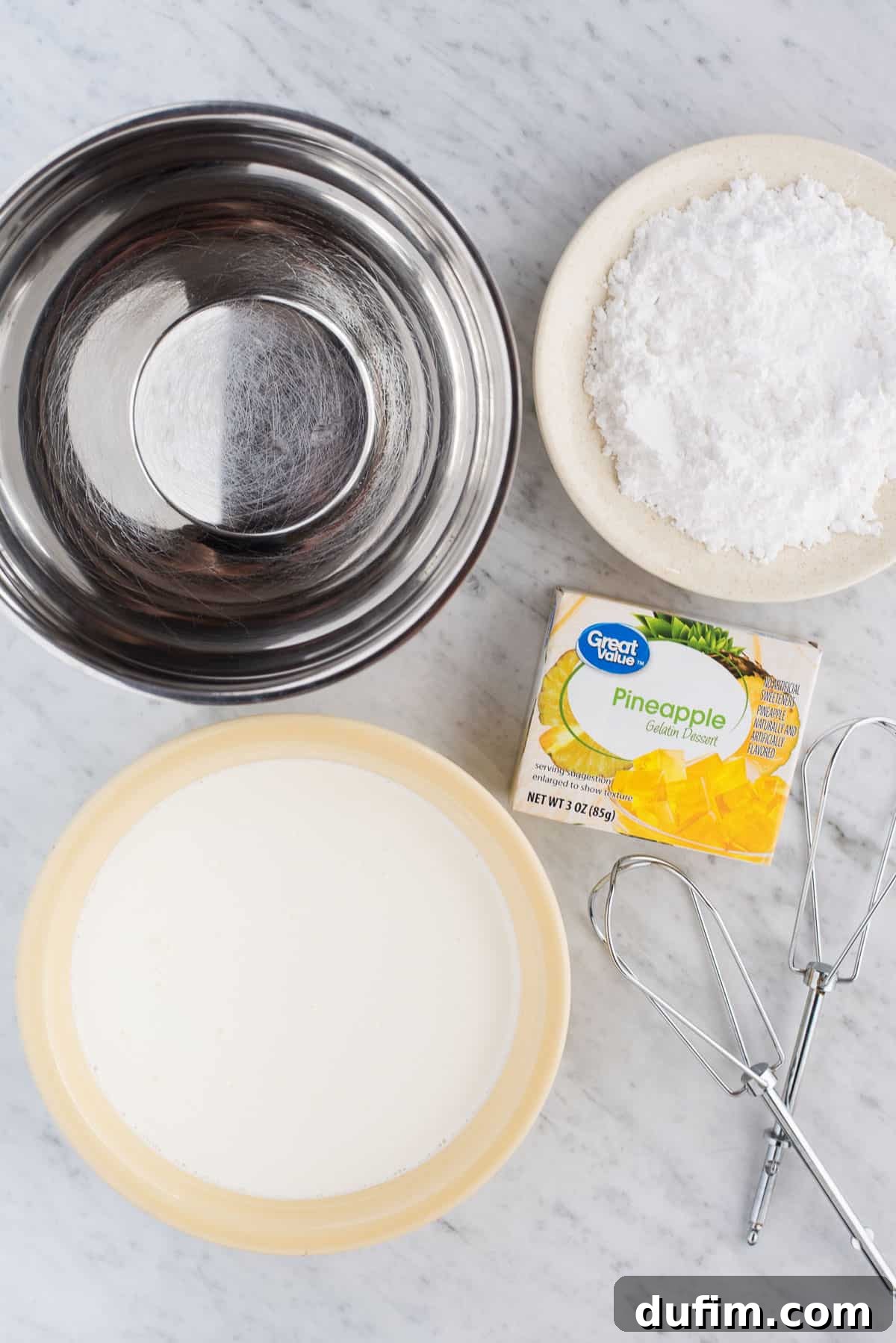 A neatly arranged flat lay showcasing the three essential ingredients for crafting perfect pineapple whipped cream: a gleaming metal mixing bowl, a bowl of rich heavy cream, a bowl of fine powdered sugar, and a vibrant box of pineapple Jell-O powder, all ready for mixing.