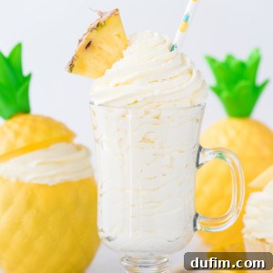 A glass mug filled with creamy pineapple whipped cream dessert, topped with a pineapple wedge and a straw with a pineapple pattern. Two decorative yellow pineapple containers and straws are in the background.