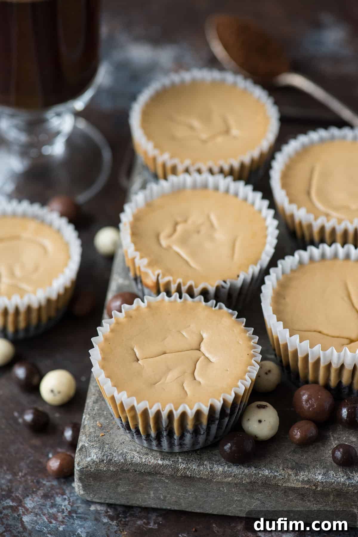 This is a crowd favorite mini coffee cheesecake recipe that you’ll use over and over! The whole recipe is only 7 ingredients and you make it in a regular size muffin pan! Plus, you can do an oreo crust or graham cracker crust for these mini espresso cheesecakes. #minicheesecake #coffeecheesecake #minicoffeecheesecake #espressocheesecake #mochacheesecake An array of freshly baked mini coffee cheesecakes, still nestled in their muffin liners, sit on a textured stone slab alongside scattered coffee beans, emphasizing their rich flavor and elegant presentation.