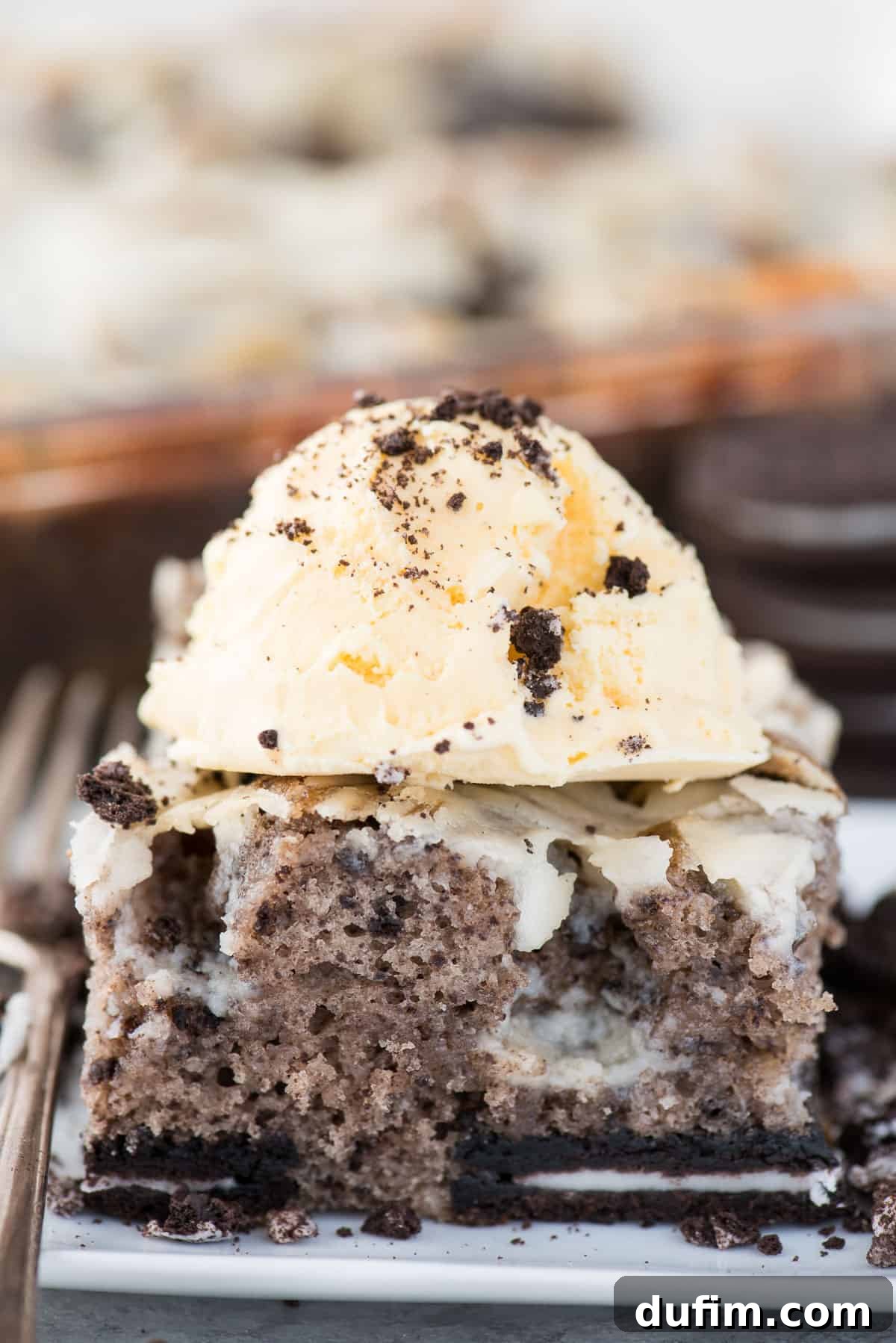 A delectable slice of Oreo Earthquake Cake, perfectly adorned with a scoop of creamy vanilla ice cream and a sprinkle of cookie crumbs, resting on a pristine white plate.
