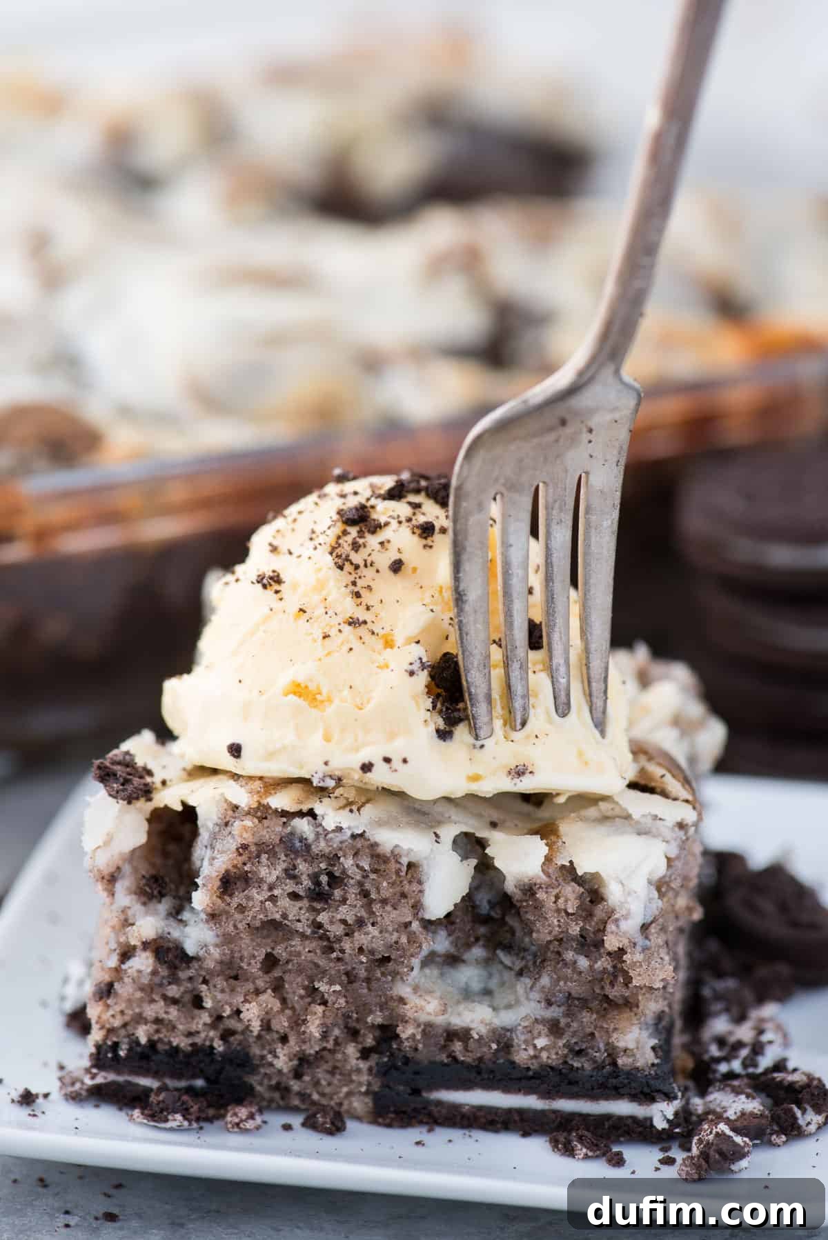 A close-up shot of a slice of Oreo Earthquake Cake, featuring the distinctive frosting crevices. A fork is inserted, ready for a taste, with vanilla ice cream and cookie crumbs on top.