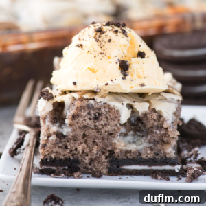 A slice of Oreo Earthquake Cake topped with a scoop of vanilla ice cream and cookie crumbs sits on a white plate, with a fork and cookie pieces scattered around.