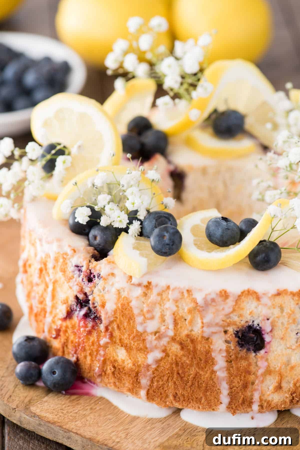 A whole lemon blueberry angel food cake, beautifully garnished with fresh lemon slices, blueberries, and baby’s breath, resting on a wooden serving plate.