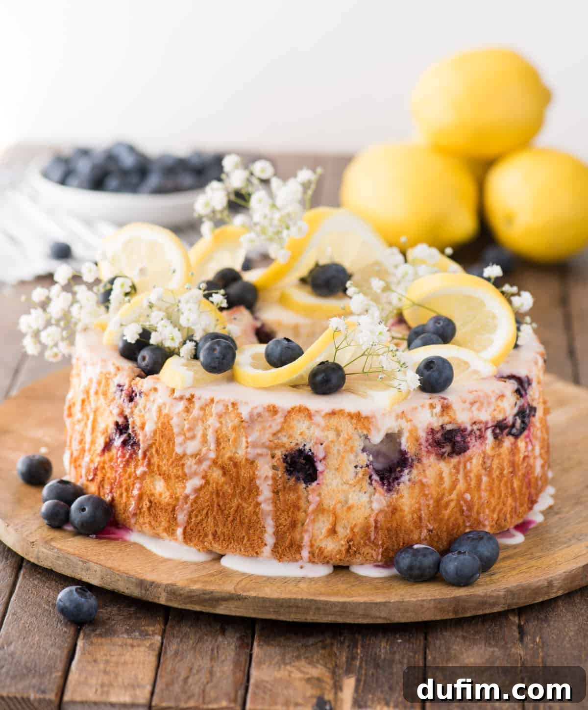 A beautiful lemon blueberry angel food cake adorned with lemon slices, fresh blueberries, and baby’s breath, ready to be served on a wooden plate.