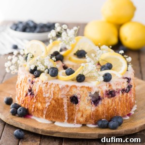 A beautifully prepared lemon blueberry angel food cake, topped with fresh lemon slices, vibrant blueberries, and delicate white flowers, sits on a rustic wooden board.
