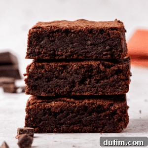 Three thick, fudgy homemade brownies are stacked on top of each other on a light surface, with chocolate chunks scattered around.