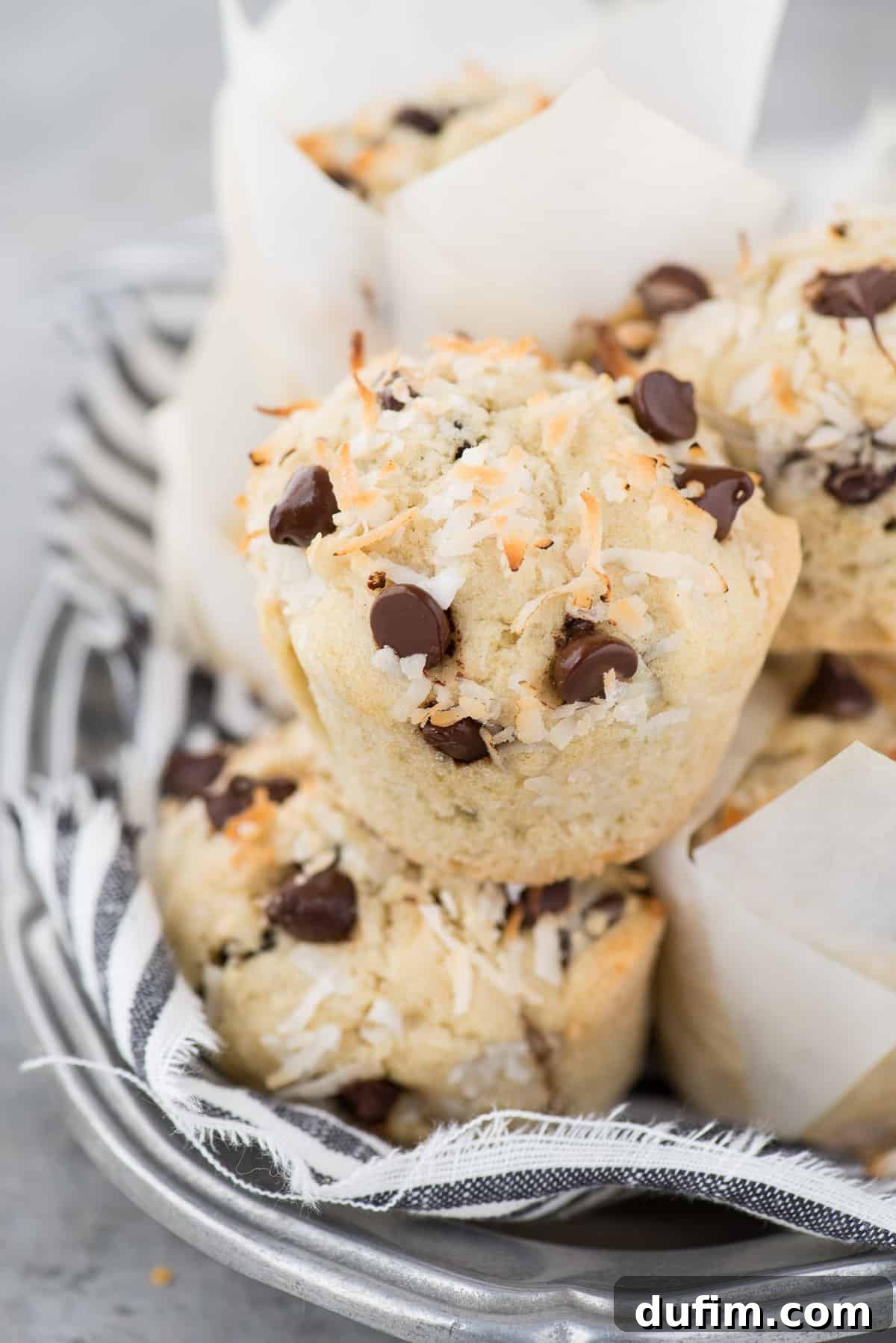 Tropical Coconut Chocolate Chip Muffins 2 Coconut chocolate chip muffins stacked in a tower on metal platter with blue and white stripe towel, showcasing their perfect domed tops and rich texture.