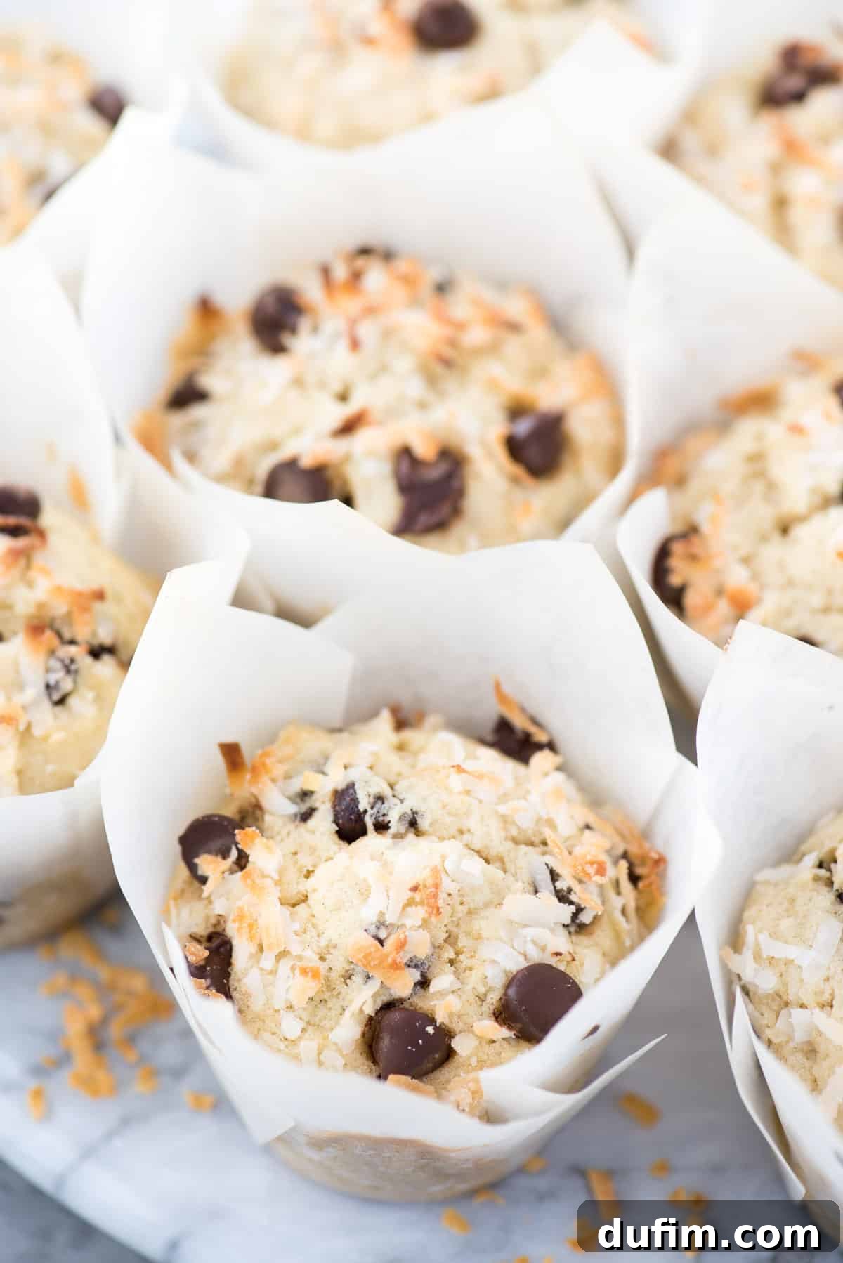 Tropical Coconut Chocolate Chip Muffins 3 Individual coconut chocolate chip muffins presented in elegant white parchment paper liners on a round marble platter, ready to be enjoyed.