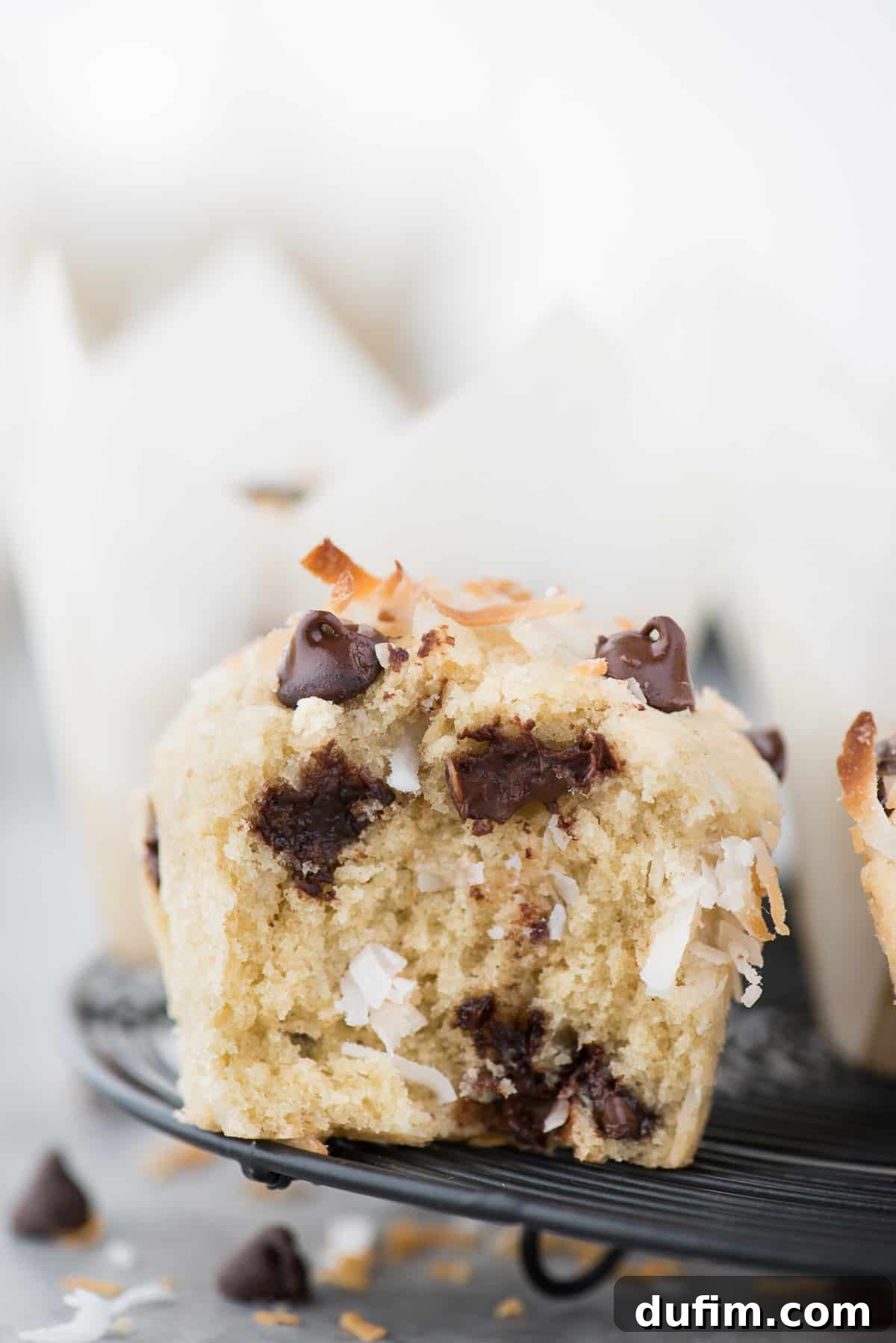 Tropical Coconut Chocolate Chip Muffins 4 A perfectly baked coconut chocolate chip muffin with a bite taken out, revealing its moist interior and generous chocolate chips, resting on a black wire cooling rack.