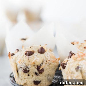 Close-up of coconut chocolate chip muffins topped with shredded coconut, resting on a black cooling rack. Chocolate chips and coconut flakes are scattered around, with more muffins blurred in the background.