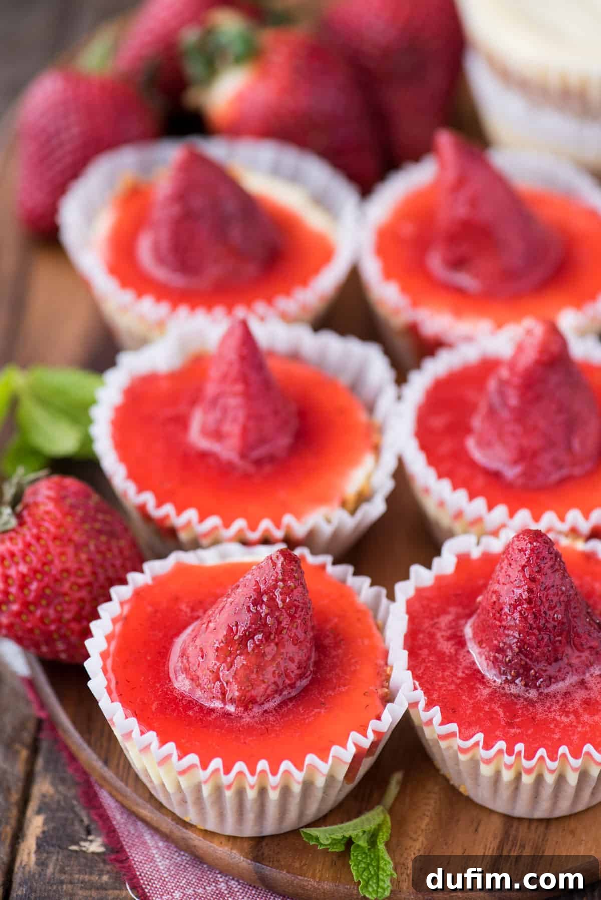 Six perfectly portioned mini plain cheesecakes adorned with a luscious strawberry topping, elegantly arranged on a rustic wooden serving tray. Fresh strawberries and mint leaves artfully garnish the presentation, inviting a taste of this delightful dessert.