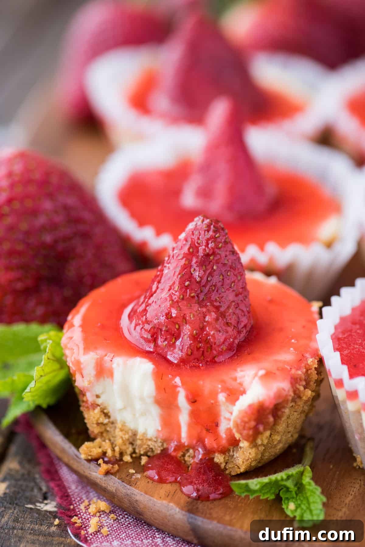 A single mini plain cheesecake, perfectly baked and boasting a creamy texture, with a spoonful of bright red strawberry topping. A bite has been removed, revealing the soft interior and crispy graham cracker crust. It rests on a wooden serving tray, suggesting a delicious treat ready to be savored.