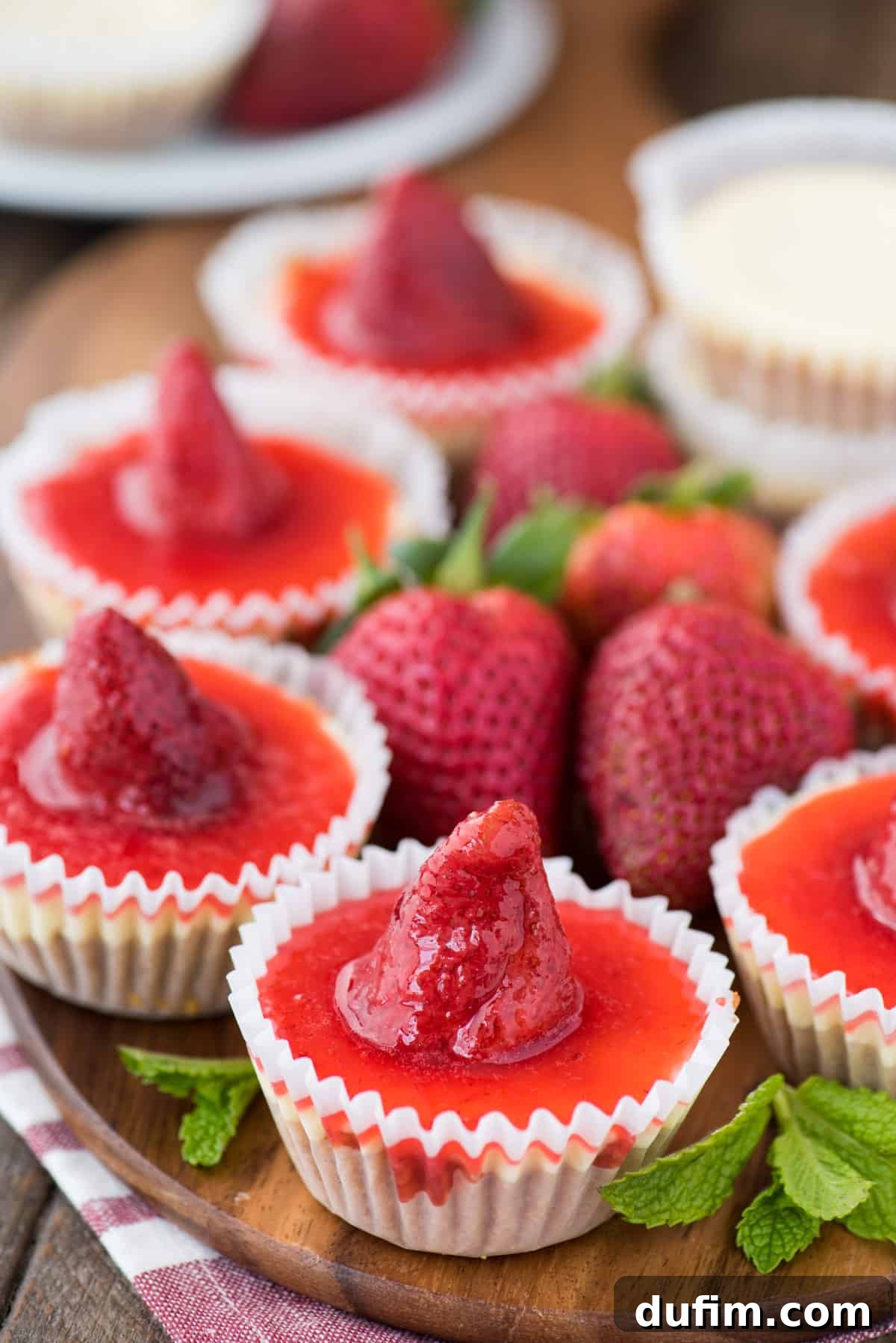 Mini plain cheesecakes with strawberry topping elegantly arranged on a rustic wooden serving tray, garnished with fresh whole strawberries and mint. The image highlights the beautiful contrast of the creamy cheesecake, vibrant red sauce, and fresh green accents.