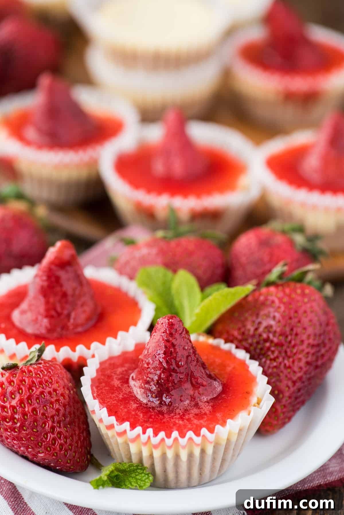 Mini plain cheesecakes with strawberry topping arranged on a white serving tray, offering a clean and elegant presentation. Fresh whole strawberries are scattered around, adding color and freshness to the dessert.