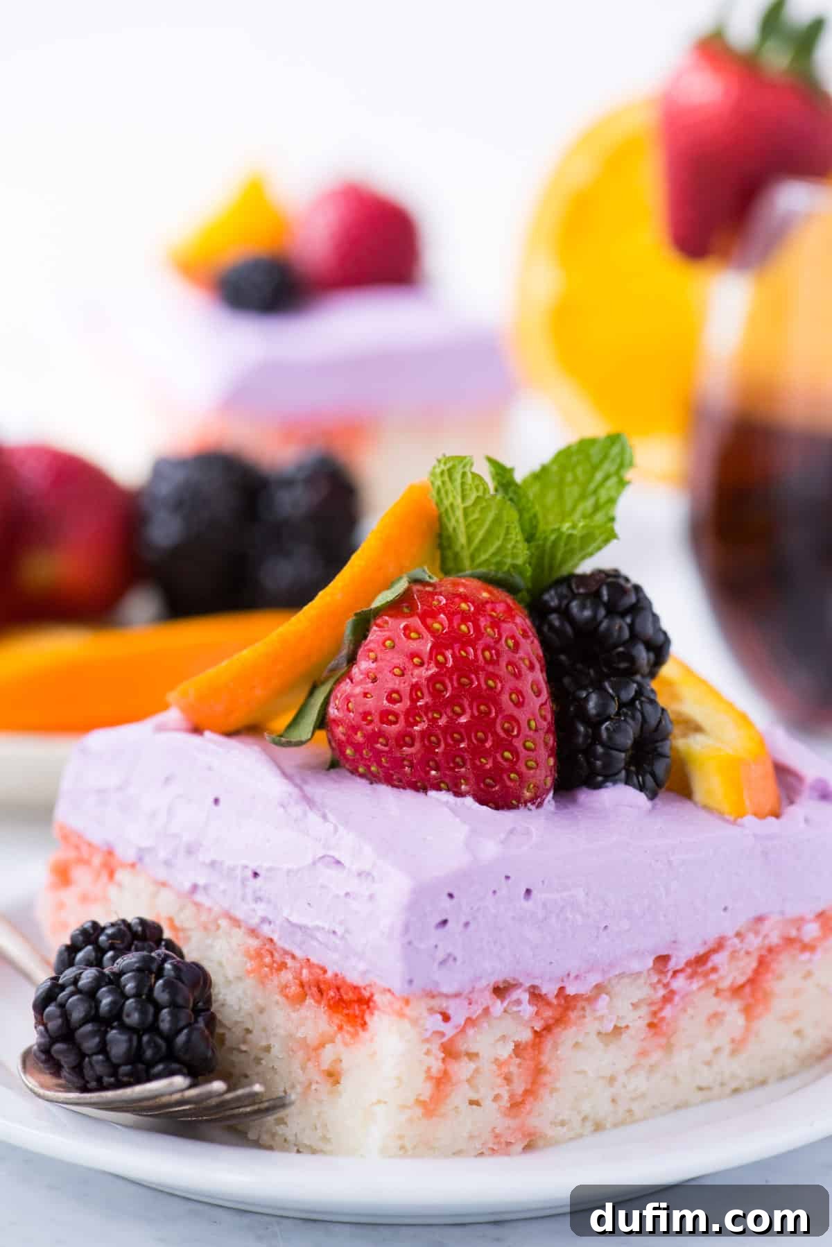 A close-up of a perfectly sliced Sangria Poke Cake, showcasing its infused, moist interior, garnished with fresh strawberries, blackberries, an orange slice, and a sprig of mint on a pristine white plate.