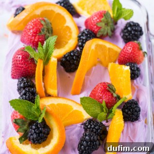 A glass baking dish filled with a vibrant purple whipped topping, elegantly garnished with slices of fresh orange, whole strawberries, juicy blackberries, and sprigs of fragrant mint, ready to be served.